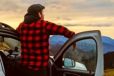 Dağcı adam, gün batımında bir macera gezisi sırasında 4x4 off-road arabasına yaslanmış dağ manzarasını seyrediyor..