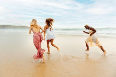 Üç genç bayanın oluşturduğu çok ırklı bir grup sahilde bir yaz gününün tadını çıkarıyor. Kumsalda koşan kızlar.