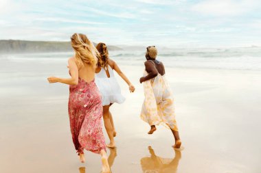 Üç genç bayanın oluşturduğu çok ırklı bir grup sahilde bir yaz gününün tadını çıkarıyor. Kumsalda koşan kızlar.