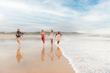 Kameraman, film yapımcısı ya da fotoğrafçı plajda koşan çok ırklı model ve aktris grubuna paralel hareket ediyor..