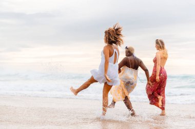 Farklı ırktan üç kız yaz tatili sırasında arkadaşlarıyla birlikte gün batımında deniz kıyısında yürüyorlar.