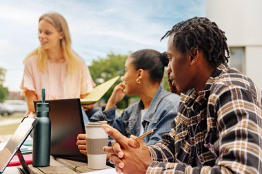 Üniversite kampüsünün dışındaki bankta oturup birlikte ders çalışan siyahi bir çocuğa odaklanan çok ırklı üniversite öğrencileri.