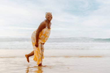Yaz elbisesi içinde genç kıvrımlı siyah kız bir yaz günü denizin kenarında yürürken gülüyor.