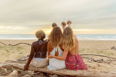 Sahilde bir kütükte oturan ve gün batımında Hindistan cevizinin içinde sunulan içecekle güneşi kızartan üç kızdan oluşan çok ırklı bir arkadaş grubu..