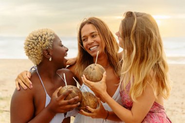 Gün batımındaki bir plaj partisinde hindistancevizinin içinde sunulan içki eşliğinde sarılıp kaynaşan üç kadının çok ırklı grup portresi. Yaz tatili