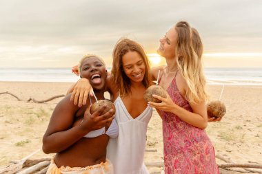 Bir yaz gezisi sırasında sahilde sunulan hindistan cevizinin tadını çıkarırken sarılıp iyi vakit geçiren üç kadın arkadaş grubu..