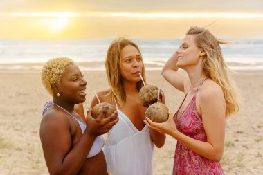 Farklı ırktan üç bayan arkadaş yaz gezisi sırasında gün batımında sahilde birlikte hindistan cevizi suyu içerler.