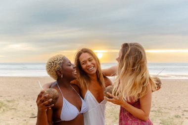 Farklı ırktan üç bayan arkadaş yaz gezisi sırasında gün batımında sahilde birlikte hindistan cevizi suyu içerler.