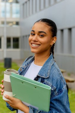 Üniversite ya da lise kampüsünün dışındaki genç Latin öğrencilerin portresi, elinde sınıf notları ve yeniden kullanılabilir fincanda kahve olan bir klasörü tutarken gülümseyen kameraya bakıyor..