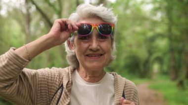 LGBT topluluğundan beyaz saçlı yaşlı bir kadının video portresi. Kameraya gülümserken gökkuşağı gurur bayrağıyla güneş gözlüğü takıyor.