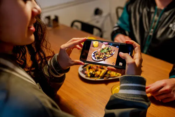 Latin kız, erkek arkadaşıyla restoranda yemek yerken akıllı telefonuyla bir tabak Meksika cipsinin fotoğrafını çekiyor. Foodie etkileyicisi sosyal ağları için içerik yaratıyor.