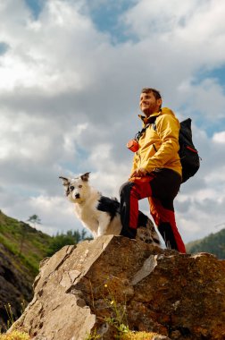 Erkek dağcı, dağ yürüyüşü sırasında sınır köpeği köpeğiyle bir kayanın üzerinde durup manzarayı seyrediyor. Spor ve macera.