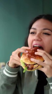 Hamburger yiyen ve kamerada mutlu görünen genç Latin kadının video portresi.