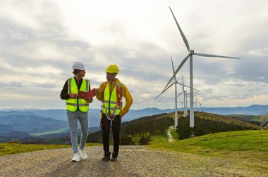 Mühendis ve operatör, sürdürülebilir enerji ve ekolojik çözümleri destekleyen bir rüzgar türbini parkında yürüme ve bakım operasyonlarını tartışıyor.