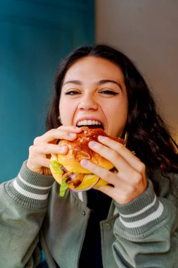 Genç bir kadın bir restoranda büyük bir lokma burger yiyor, yemeğin tadını çıkarıyor.