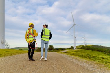 Mühendis ve operatör rüzgar türbini parkında yürüyor ve konuşuyor, yenilenebilir enerji ve bakımı tartışıyorlar.