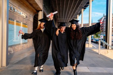 Bir grup neşeli, çok kültürlü mezunlar. Önlük ve havan mermisi giymiş. Sokakta koşuyorlar ve diplomalarını tutuyorlar.