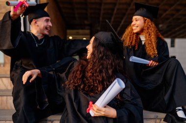 Bir grup neşeli çoklu etnik mezun önlük ve havan mermisi giymiş, ellerinde diplomalar ve mezuniyetlerini kutluyorlar.