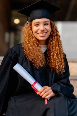 Diplomasını elinde tutan, mezuniyet elbisesi giyen ve gülümseyen neşeli bir üniversite öğrencisinin portresi.