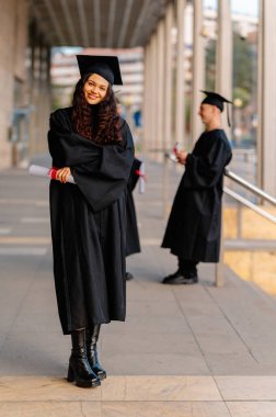 Mezun olan genç bir kadın, mutlu bir mezuniyet gününde diplomasını alıp kollarını kavuşturarak gülümsüyor.