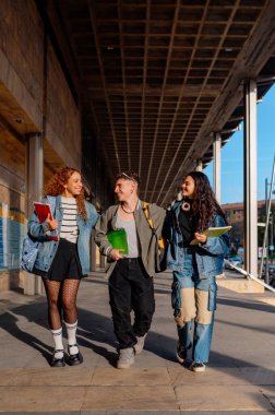 Modern bir üniversite kampüsünde birlikte yürüyüp konuşan üç neşeli öğrenci ders aralarında eğleniyorlar.