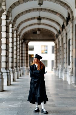 Üniversitesinde kemerin altında duran, diplomasını tutan ve akademik başarısını kutlayan kendinden emin mezun.