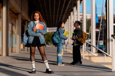 Arkadaşları arka planda sohbet ederken bir üniversite kampüsünde notlarla poz veren genç bir kadın.