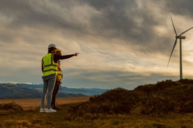 Mühendisler, gün batımında dağlık arazide rüzgar türbini kurulumu projesinin sonraki adımlarını tartışıyorlar.
