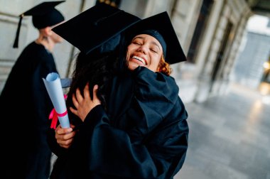 İki mutlu kadın mezun üniversite koridorunda birbirlerine sarılıp mezuniyetlerini kutluyorlar.