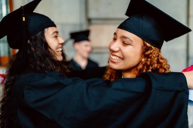 Havan topu takan iki gülümseyen etnik kadın mezun mezuniyet törenlerinde birbirlerine sarılıyorlar.