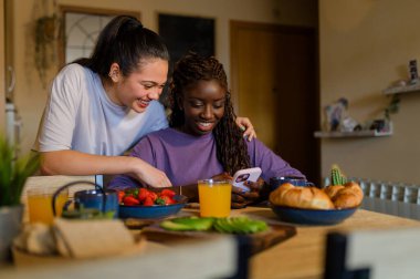 Mutlu ırklar arası lezbiyen çift evde keyifli bir kahvaltı yapıyor, sıcak bir ortamda taze meyve ve hamur işleriyle kaynaşıyorlar.
