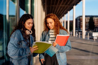 Kampüste birlikte oturan iki genç Latin kadın ders notlarını gözden geçiriyorlar ve bir sonraki derslerine katılmadan önce çalışma materyallerini tartışıyorlar.