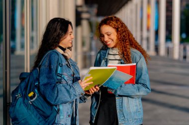 İki neşeli kız öğrenci üniversite binasının önünde oturmuş notlarını tartışıyorlar ve güneşli bir günün tadını çıkarıyorlar.