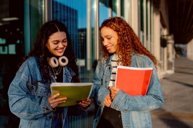 İki neşeli çok ırklı kız öğrenci bir üniversite binasının dışında notlarını karşılaştırıyor ve fikirlerini paylaşıyor, birlikte akademik yolculuklarını kutluyorlar.