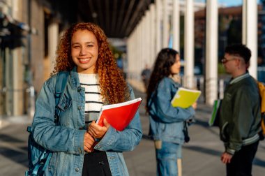 Elinde kitaplarla kampüste gülümseyen neşeli bir üniversite öğrencisinin portresi.
