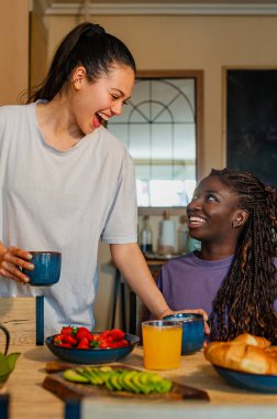 Mutlu lezbiyen çift gülüyor ve evde çilek, avokado, kruvasan, portakal suyu ve kahveyle sağlıklı bir kahvaltı yapıyor.