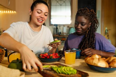 İki bayan ev arkadaşı evlerinin mutfağında meyve, avokado tostu ve portakal suyuyla sağlıklı bir kahvaltı yapıyorlar.