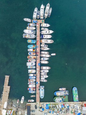 Hareketli bir marinanın yüksek açılı görüntüsü, çeşitli teknelerin sergilendiği, yatlardan küçük balıkçı teknelerine kadar, düzgün bir şekilde kenetlenmiş. 