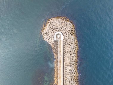 Deniz feneriyle birlikte bir kıyı dalgasının havadan görüntüsü. Yüksek açılı, turkuaz suya doğru uzanan bir dalgakıranın tam görüntüsü, geniş, açık bej beton bloklar dizisi bir distin oluşturur.