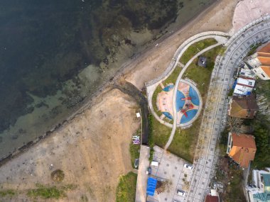 Oyun parkı ve konut binaları olan Coastal Park 'ın havadan görüntüsü. Yüksek açılı, canlı bir oyun parkı, kıvrımlı yürüyüş yolları ve yerleşim yerine giden asfalt bir yol olan bir sahil parkı.