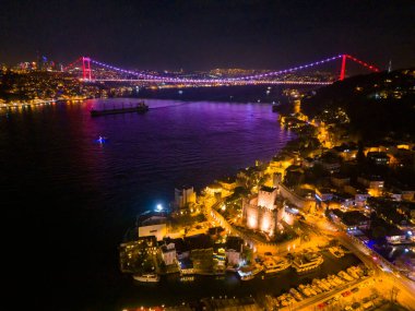 İstanbul 'un Gece Boğazı: Aydınlatılmış Köprü ve Kale. İstanbul Boğazı 'nın gece hava manzarası aydınlanmış Boğaz Köprüsü ve tarihi Anadolu Kalesi' ni gözler önüne seriyor.