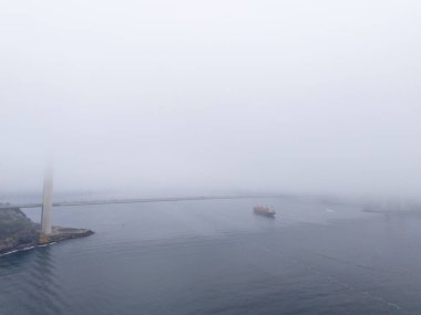 Gemisi ve deniz feneri olan Misty Harbor View. Sisli bir limanın hava görüntüsü, uzun bir deniz feneri ve birkaç gemi sergileniyor. Yavuz Sultan Selim Köprüsü İstanbul Türkiye Beykoz Poyrazkoy Anadolu