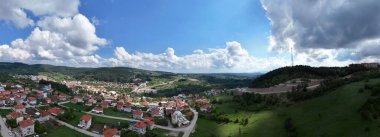 Bursa, Türkiye 'de küçük bir kasaba olan Keles' in panoramik hava manzarası. Kırmızı çatılı evler, yemyeşil tepeler ve dramatik bulutlar geniş mavi gökyüzü altında manzaralı kırsal bir manzara yaratır.
