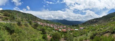 Türkiye 'nin Keles Kemaliye köyünün panoramik manzarası, yeşil tepeler, geleneksel evler ve dramatik bulutlu gökyüzü, bölgenin barışçıl kırsal yaşamını ve doğal güzelliğini yansıtıyor.