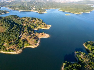 Bereketli yeşil ormanlarla çevrili sakin mavi suların ve berrak bir gökyüzü altında yuvarlanan tepelerin bulunduğu Merli Barajı 'nın havadan görünüşü manzaranın dingin ve doğal güzelliğini yansıtıyor..