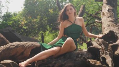 Bir bitkinin dikenli köklerini tutan genç bir kadın mayoyla ağaç kökleri üzerinde oturuyor. Yüksek kalite 4k video kaydı