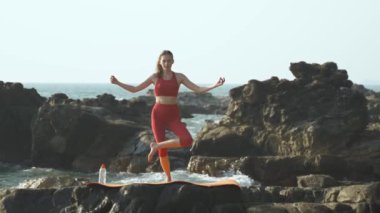 Genç bir kadın yoga pozunda tek bacağının üzerinde dengede duruyor, arkasında da okyanus taşları var. Yüksek kalite 4k video kaydı