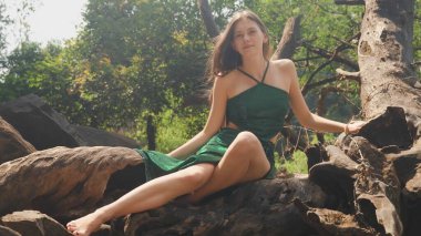 Bir bitkinin dikenli köklerini tutan genç bir kadın mayoyla ağaç kökleri üzerinde oturuyor. Yüksek kalite fotoğraf