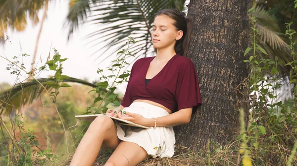 young woman sits under a palm tree, reading a book. High quality photo