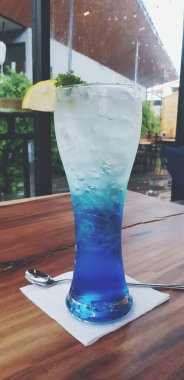 Taze mavi Hawaii İtalyan soda kokteyli, limon dilimleri ve ahşap masanın üzerindeki yaprakların üzerinde, kafenin arka planında, klasik renk tarzında. İçecek bir şeyler.